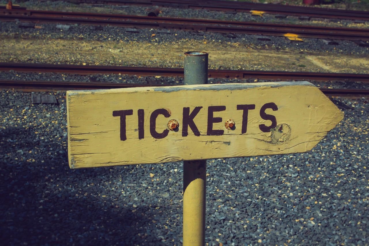 Weathered wooden ticket sign pointing towards railway tracks outdoors, vintage look.